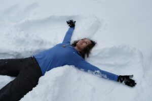A joyful woman in blue enjoying winter by making a snow angel outside.